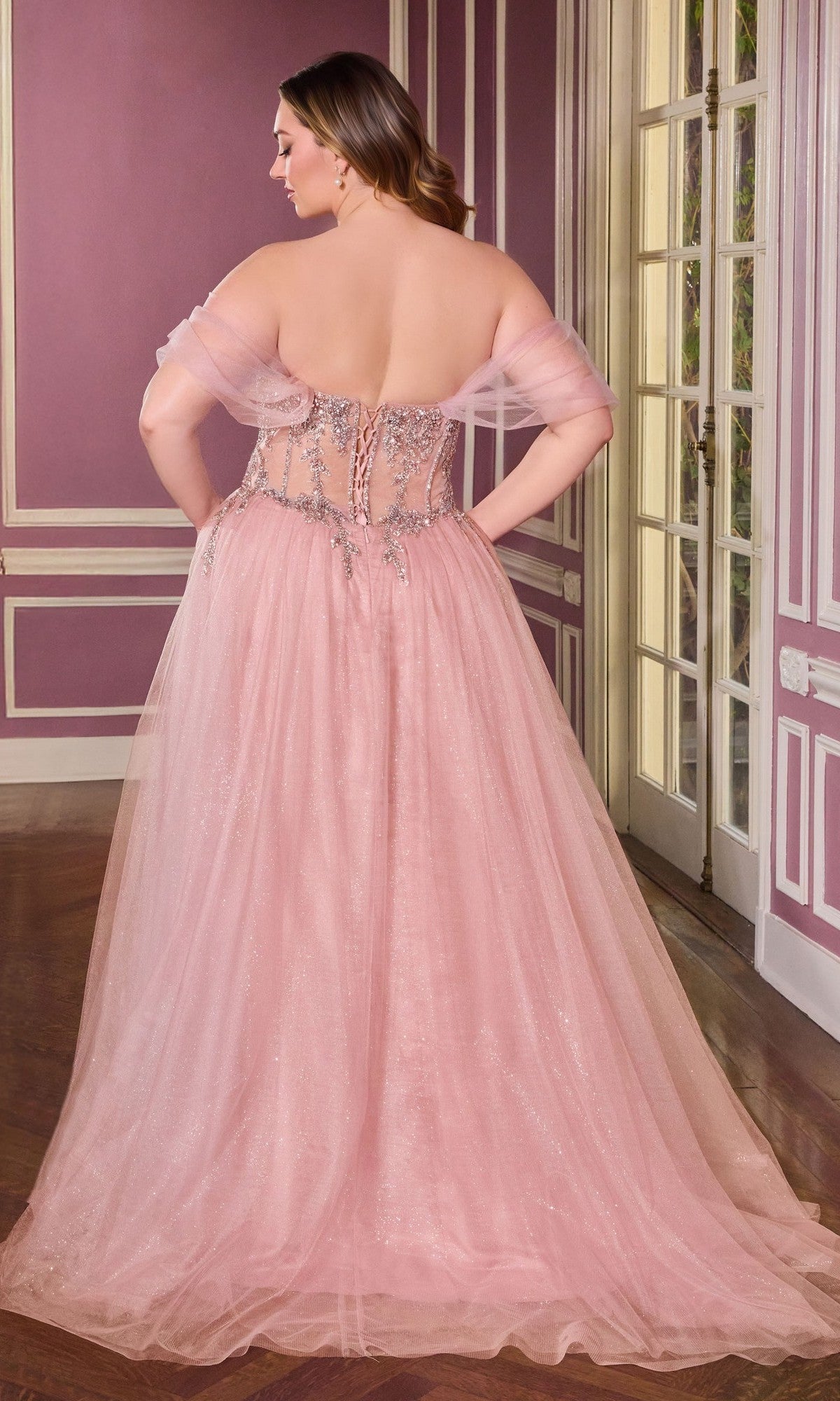 A woman stands indoors facing away, wearing the Long Plus-Size Prom Dress Ladivine CD721C—a pink glitter tulle ball gown with an embellished corset bodice and flowing skirt.