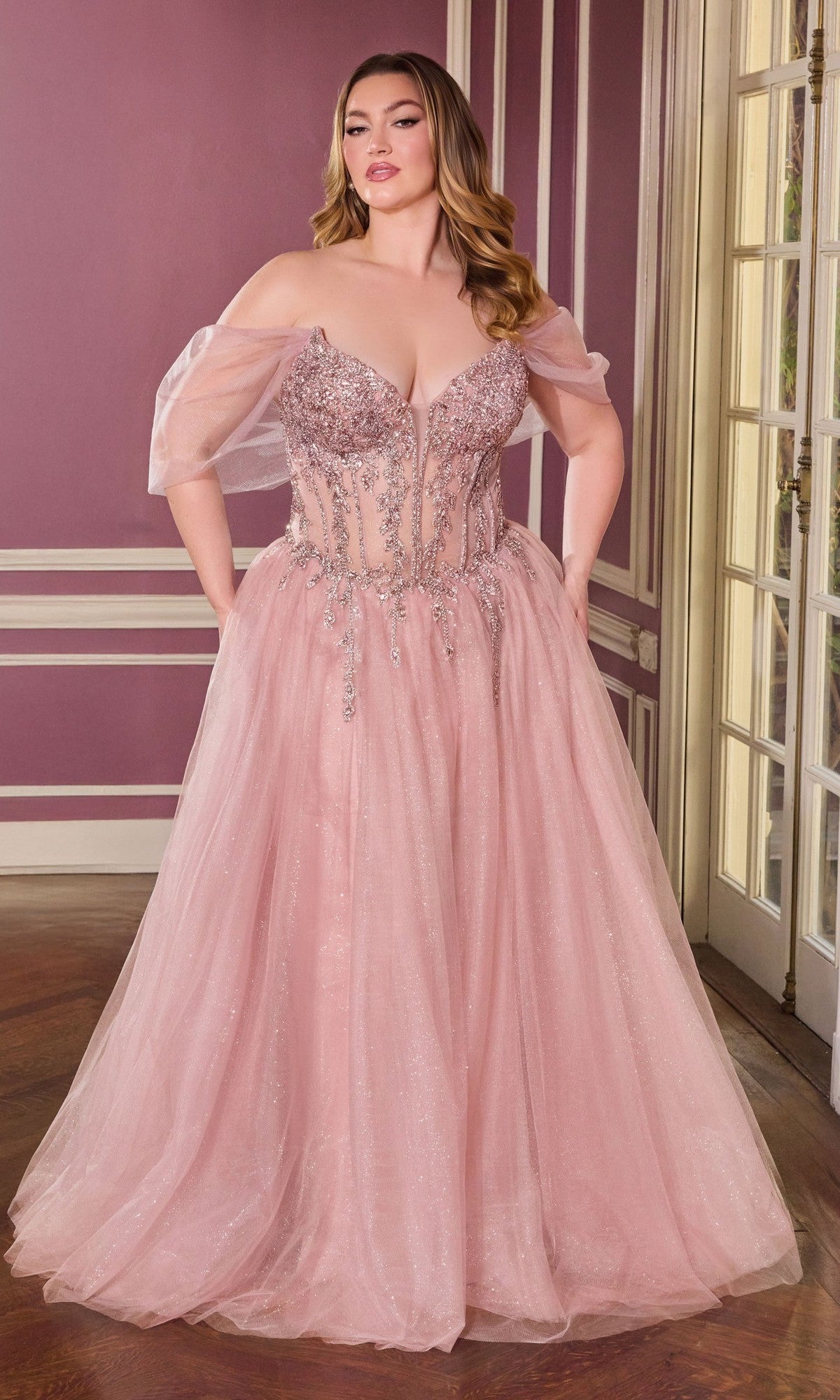A woman stands indoors wearing the Ladivine CD721C, a long, plus-size mauve prom dress featuring an embellished corset bodice and sheer tulle off-the-shoulder sleeves.
