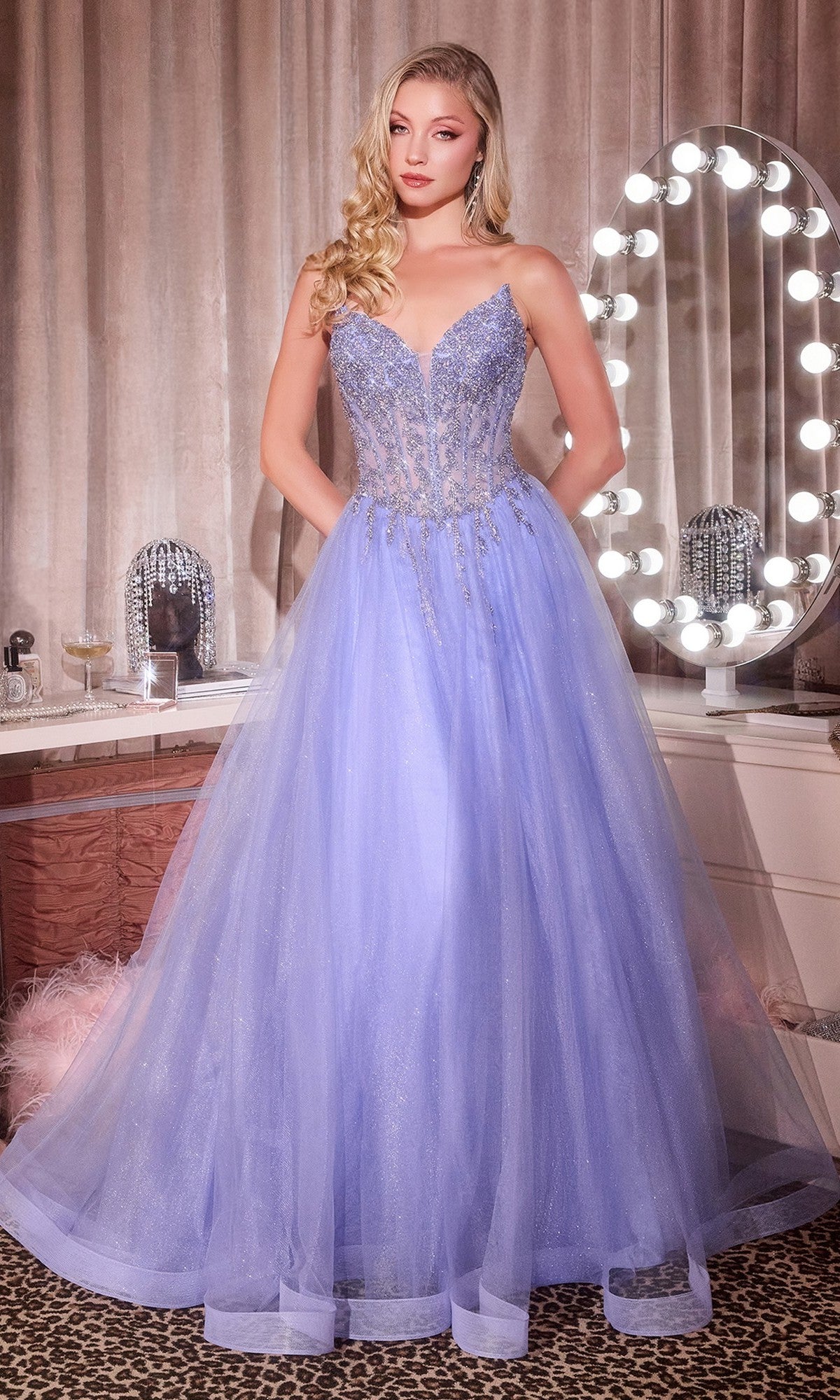 A woman in the Lace-Up Strapless Prom Ball Gown: Ladivine CD721, a light purple glitter tulle A-line dress, stands by a vanity mirror under soft lighting.