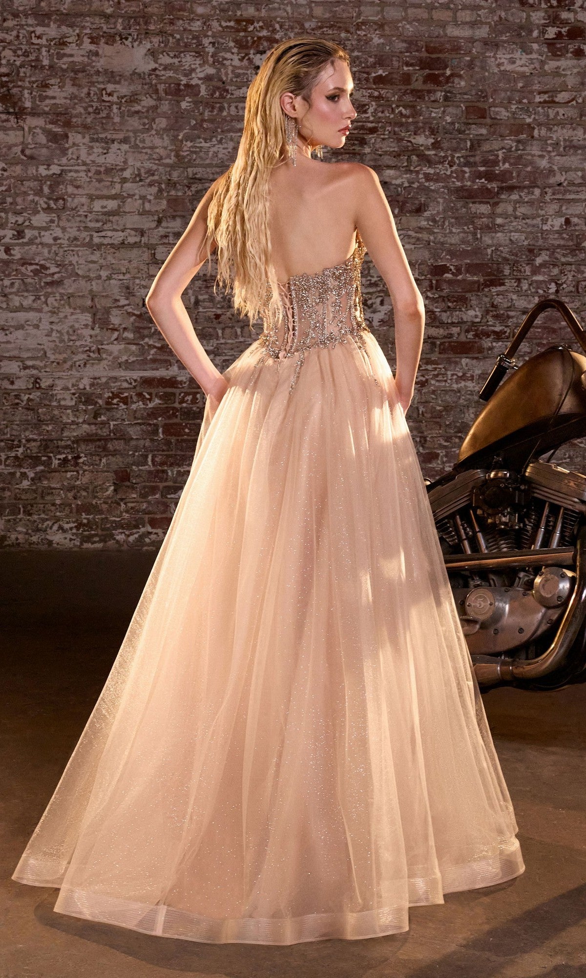 A woman in the Lace-Up Strapless Prom Ball Gown: Ladivine CD721 stands before a brick wall and motorcycle, facing away to showcase the beaded corset and long a-line silhouette.