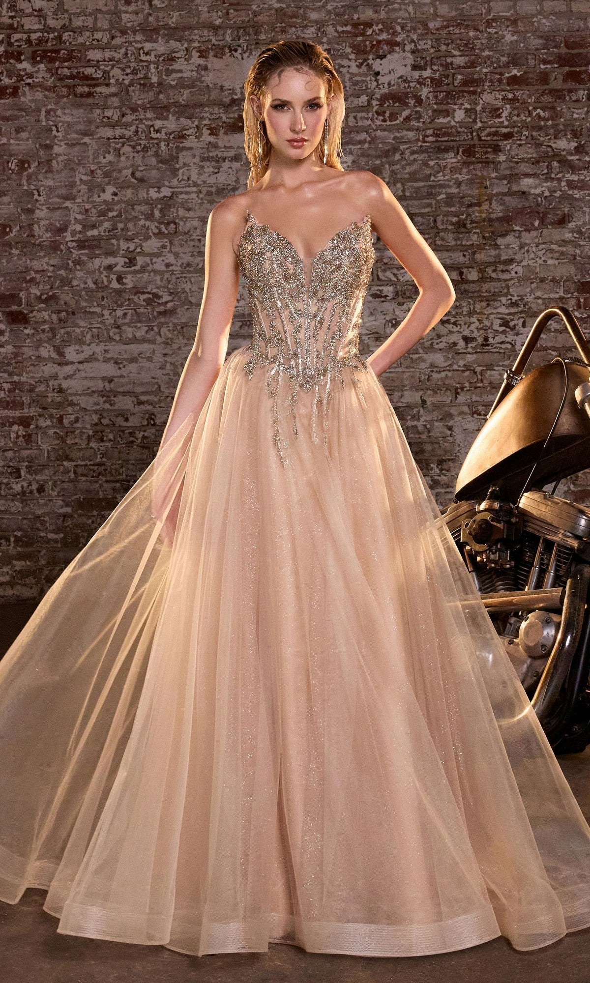 A woman in the Lace-Up Strapless Prom Ball Gown: Ladivine CD721, featuring a beaded corset and glitter tulle, stands beside a motorcycle against a brick wall.