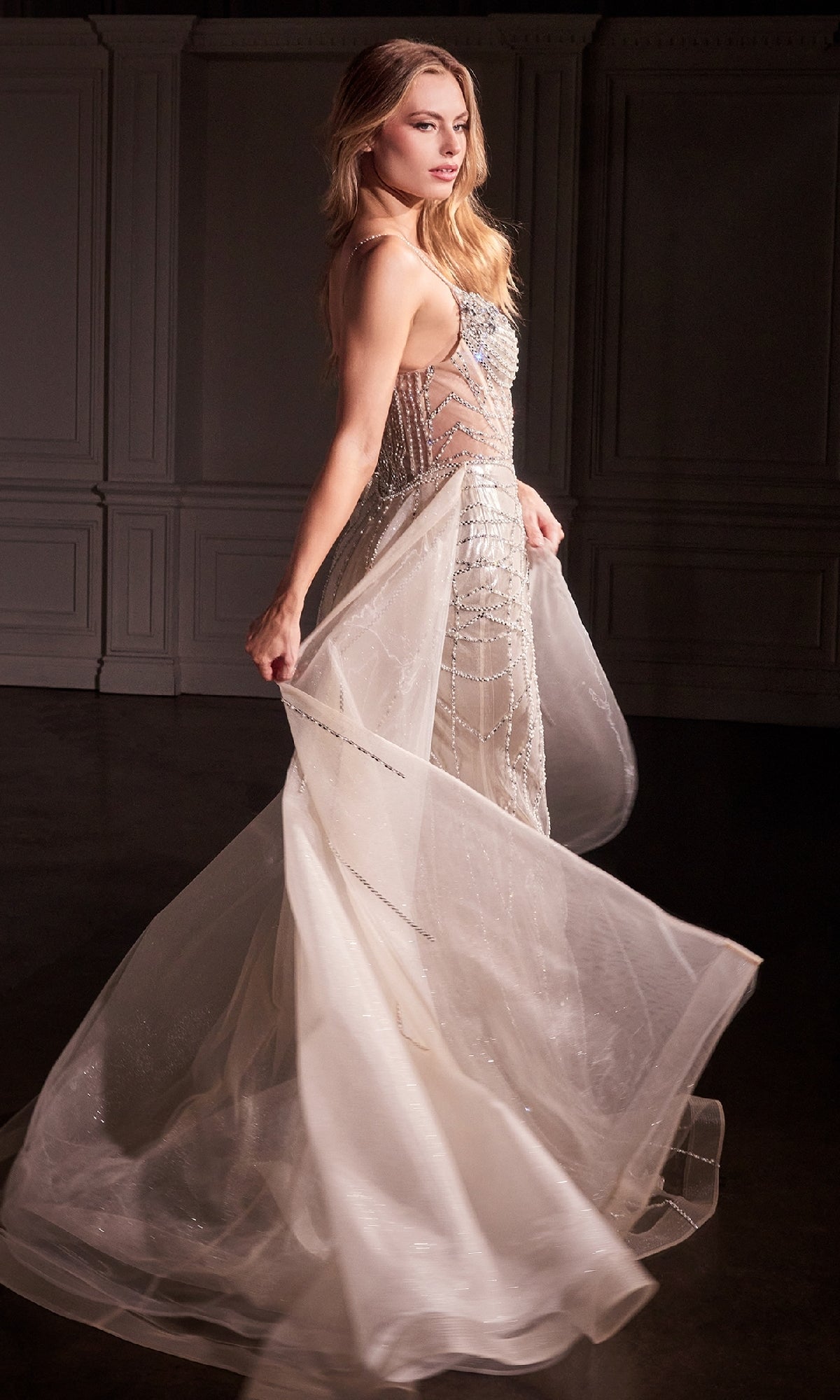 A woman in the Ladivine CD763 Overskirt Long Glitter Prom Dress, featuring a sleeveless sheer white design with intricate beadwork and a removable overskirt, stands in a dimly lit room with white paneled walls, gently holding the dress.