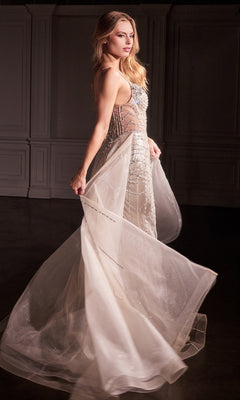 A woman in the Ladivine CD763 Overskirt Long Glitter Prom Dress, featuring a sleeveless sheer white design with intricate beadwork and a removable overskirt, stands in a dimly lit room with white paneled walls, gently holding the dress.