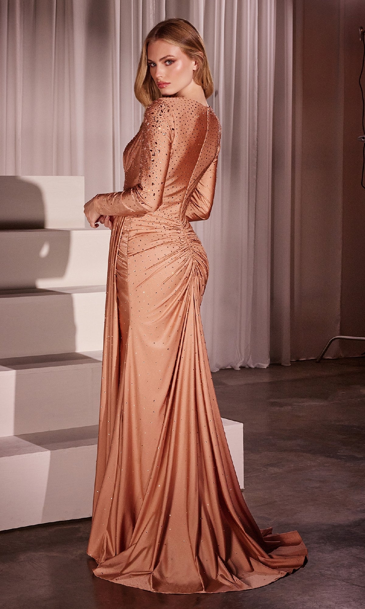 A woman stands in front of white drapes wearing the CLCD788 Long Sleeve Long Beaded Prom Dress, a fitted copper evening gown with beaded embellishments and gathered detailing at the back.