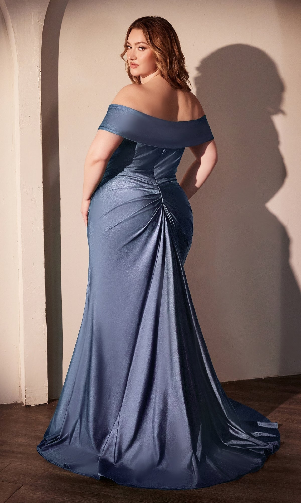 A woman in the Ladivine CD790C Off-Shoulder Plus-Size Prom Gown, a shimmery blue evening dress under $150, stands with her back to the camera against a white wall, casting an elegant shadow.