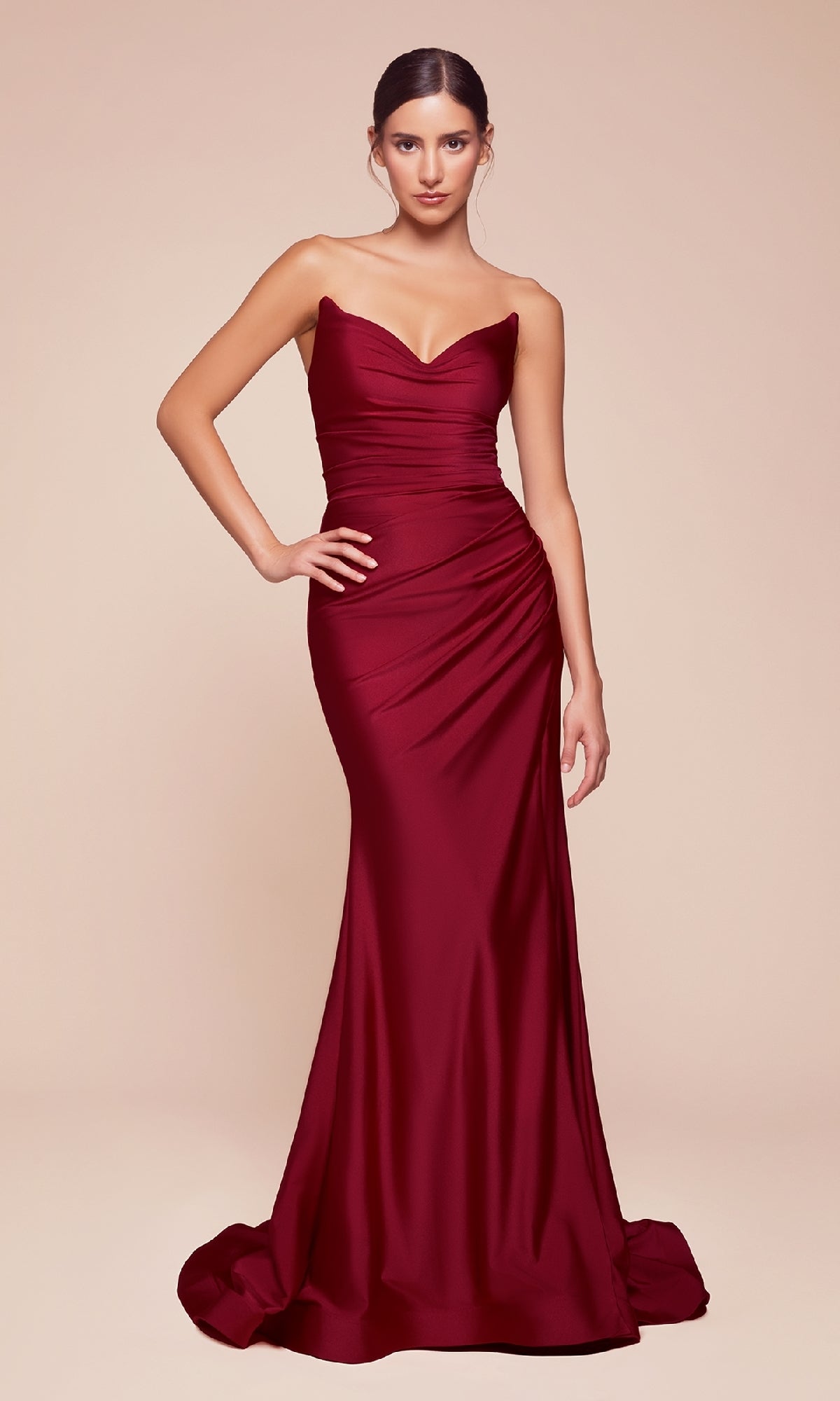 A woman stands against a beige background wearing the Lace-Up Strapless Long Prom Dress: Ladivine CD791 in burgundy, featuring booty-contouring ruching and a floor-length silhouette with a slight train.