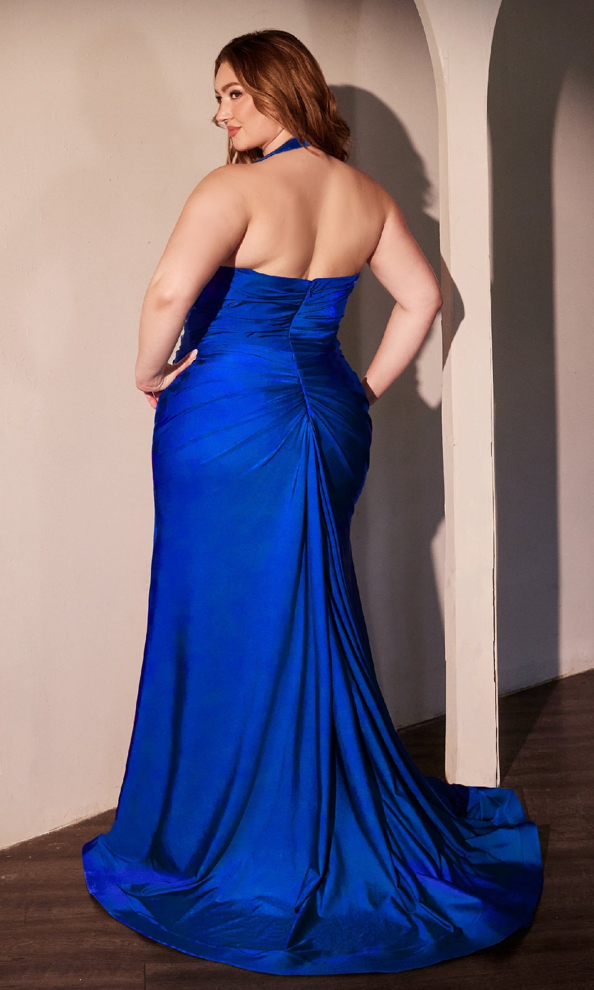 A woman in the Plus-Size Long Halter Prom Dress: Ladivine CD796C, a blue floor-length gown, stands with her back to the camera and one hand on her hip beside a white archway.