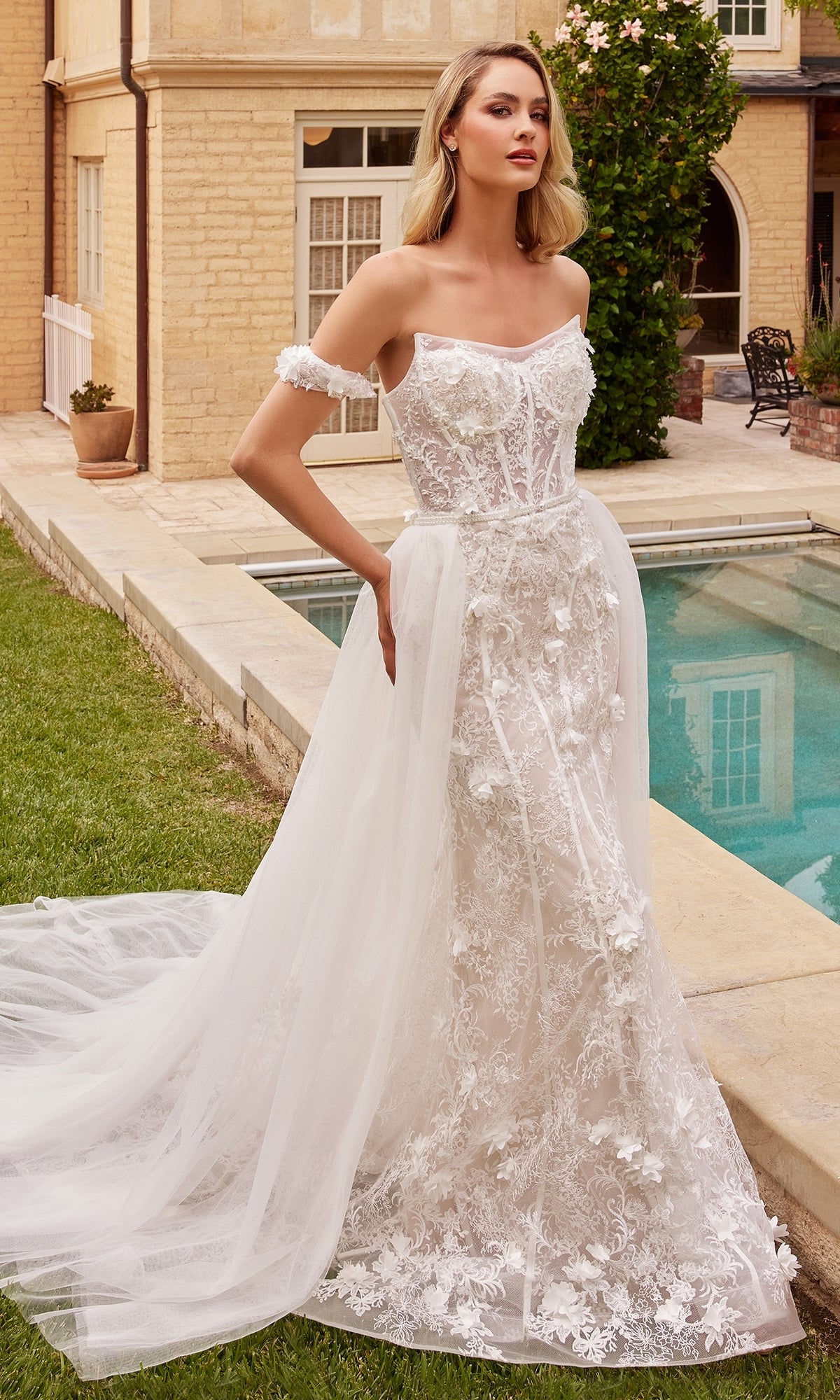 A woman stands by a pool in the 3D Floral Lace Wedding Gown CD861W—an off-shoulder, off-white dress with floral appliqué and a removable tulle overskirt, perfect for an elegant outdoor bridal look.