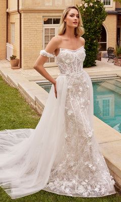A woman stands by a pool in the 3D Floral Lace Wedding Gown CD861W—an off-shoulder, off-white dress with floral appliqué and a removable tulle overskirt, perfect for an elegant outdoor bridal look.