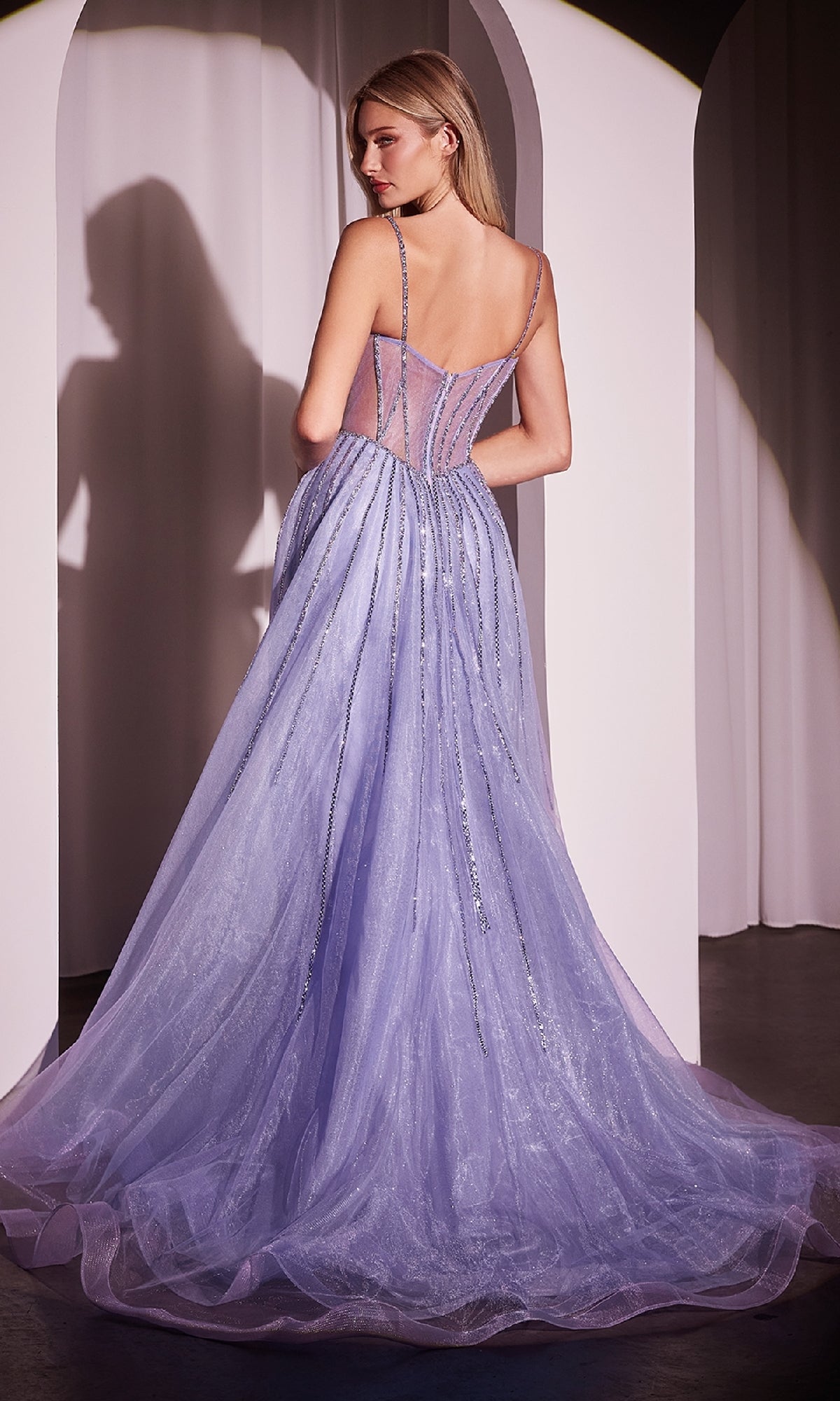 Woman in the Beaded Overskirt Long Formal Prom Dress CD863—a lavender, floor-length gown with spaghetti straps and a fitted semi-sheer bodice—stands with her back to the camera, highlighting its elegant, prom-inspired design.