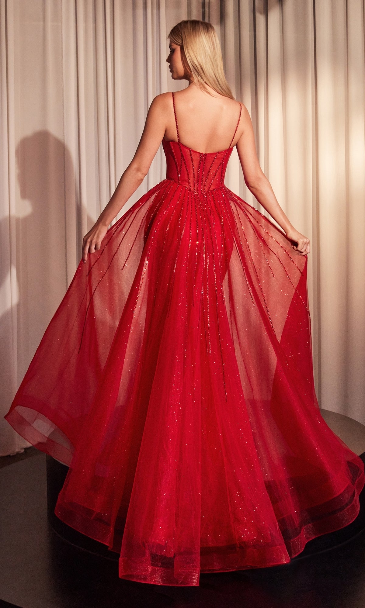 A woman stands facing away, holding the sides of her Beaded Overskirt Long Formal Prom Dress CD863—a red, floor-length formal gown with a sheer, sparkling overlay. White curtains are in the background.