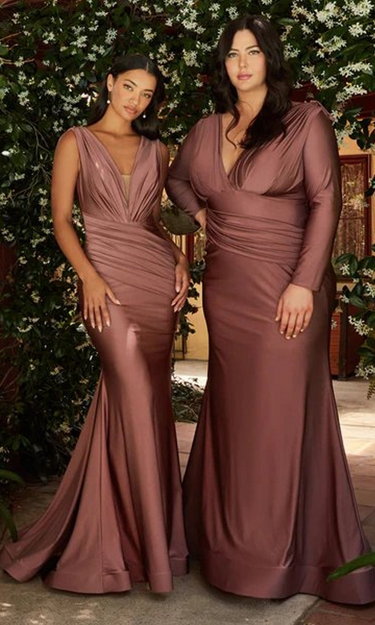 Two women stand side by side wearing the Pleated-Waist Long Sleeveless Prom Dress CD912, showcasing its curve-enhancing silhouette and draped details, posed before green foliage and white blooms.