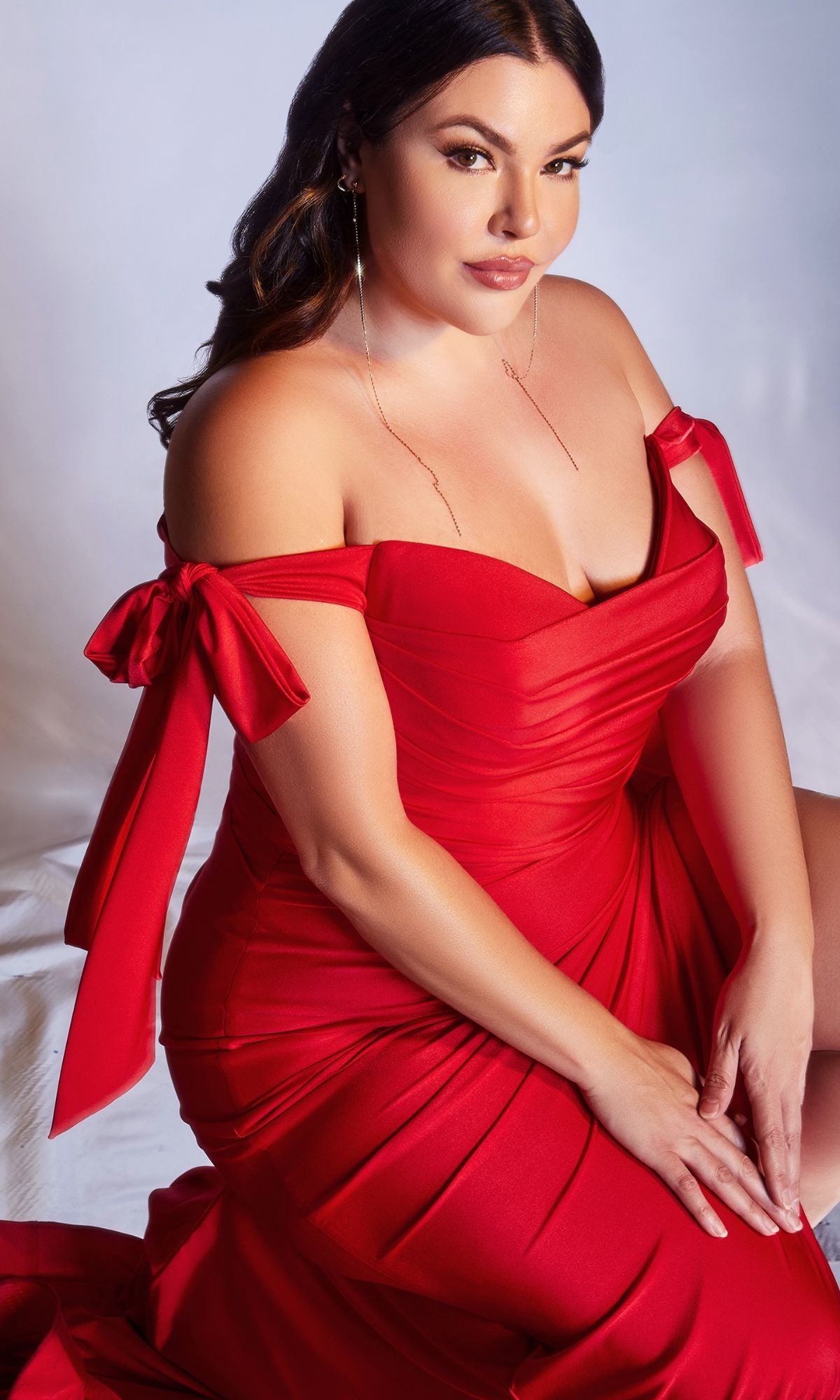 Woman with long brown hair wears the Plus-Size Long Prom Dress with Bows CD943C, sitting on a white surface and looking at the camera.