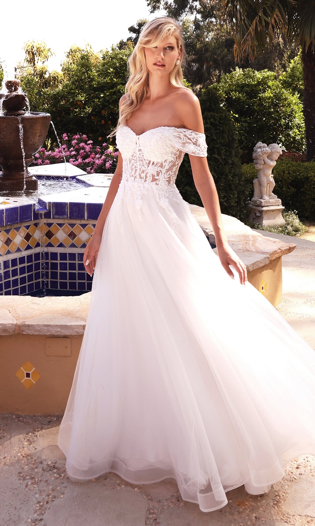 A woman stands outdoors near a tiled fountain and garden, wearing the Sheer-Bodice Long White Prom Ball Gown CD961W featuring an off-the-shoulder lace bodice and flowing tulle skirt.