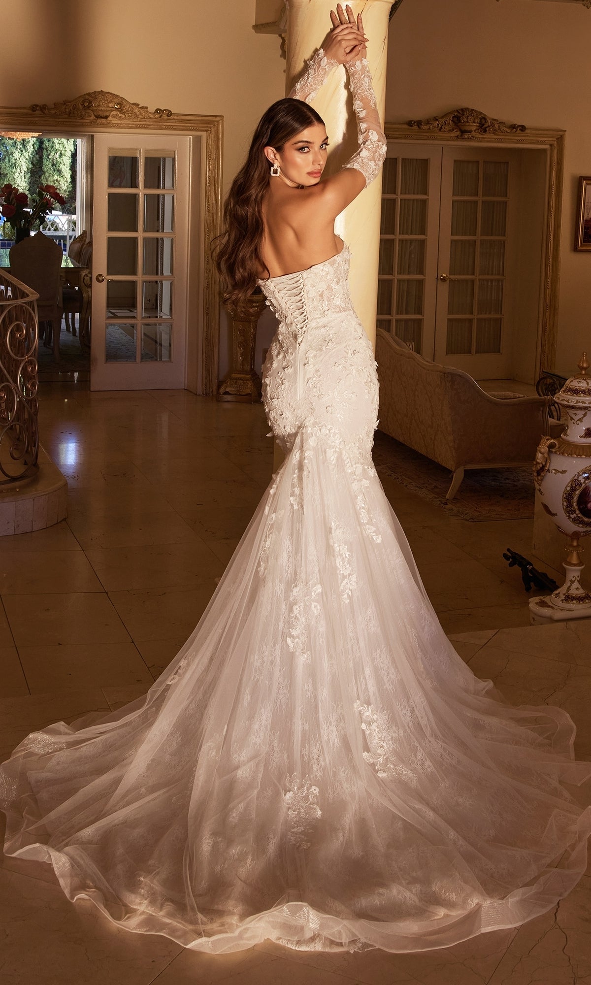 A woman stands indoors, looking over her shoulder, wearing the Long White Strapless Bridal Gown CD977W featuring a mermaid skirt and embroidered floral designs.
