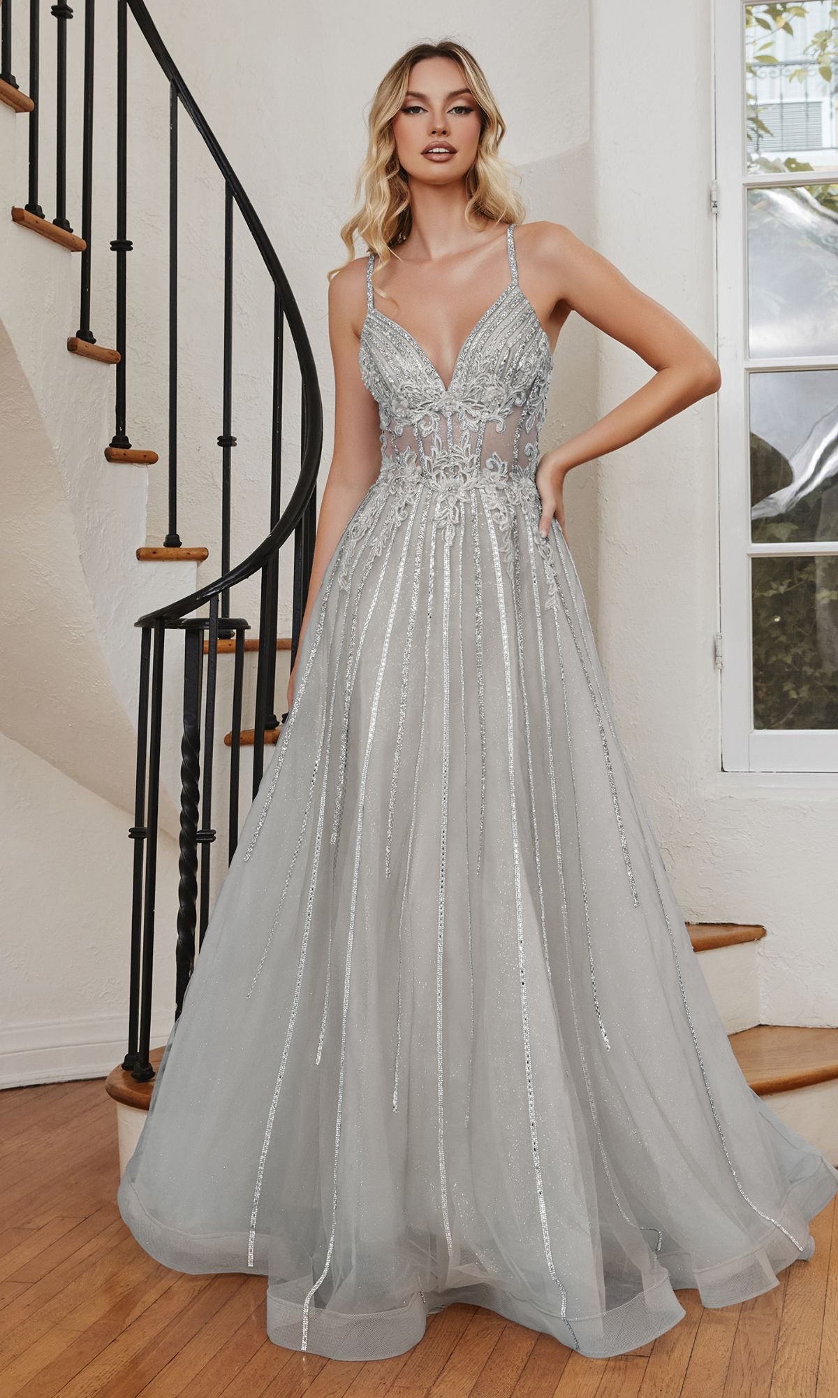 A woman stands indoors by a spiral staircase, wearing the Long A-Line Prom Dress with Sheer Bodice CD994 featuring silver sequins and a flowing tulle skirt.