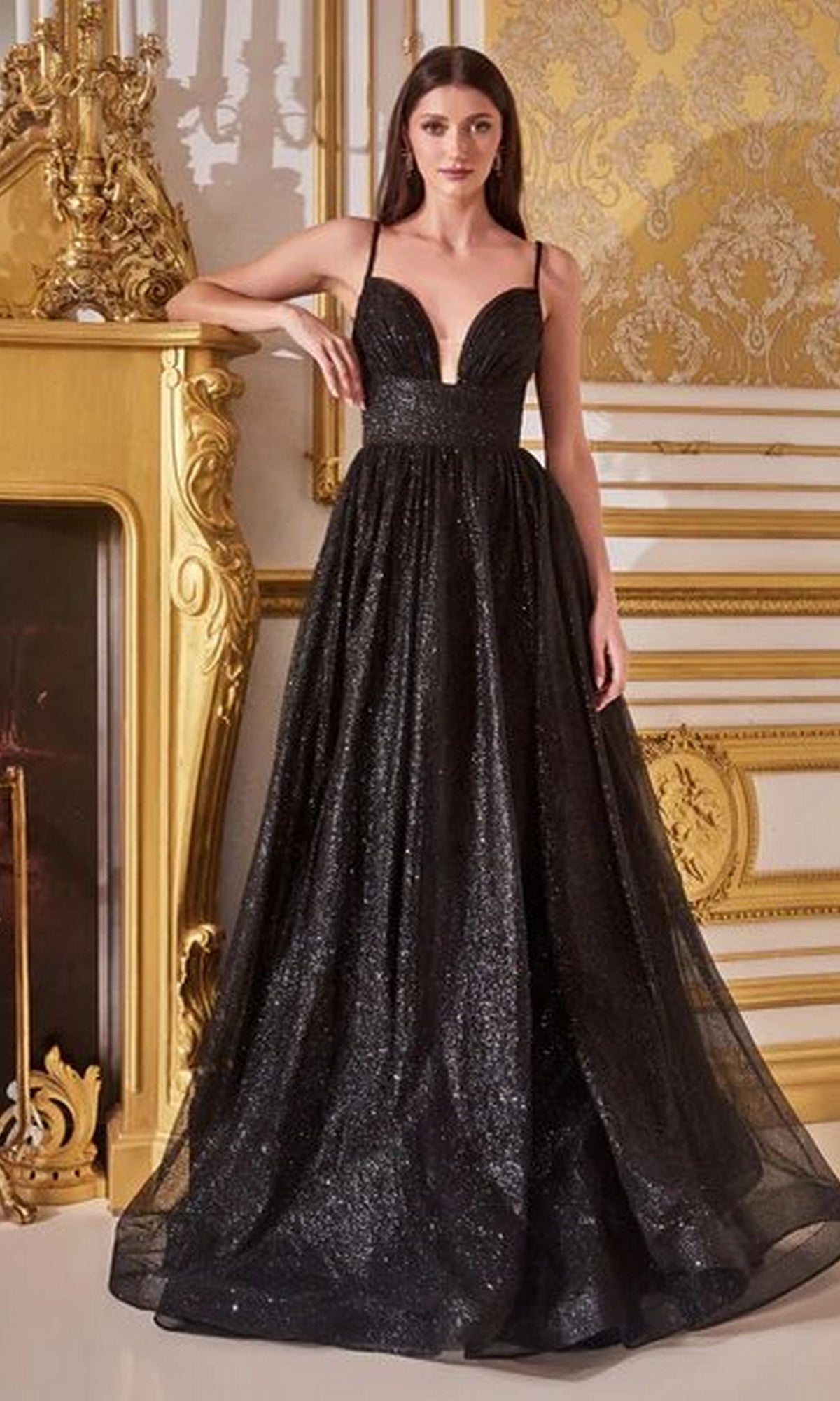 A woman stands indoors wearing the Corset-Back Long Glitter Prom Ball Gown CD996, featuring a sleek, sleeveless design and deep V neckline. Ornate gold decor and a fireplace in the background enhance the gown’s elegant appeal.