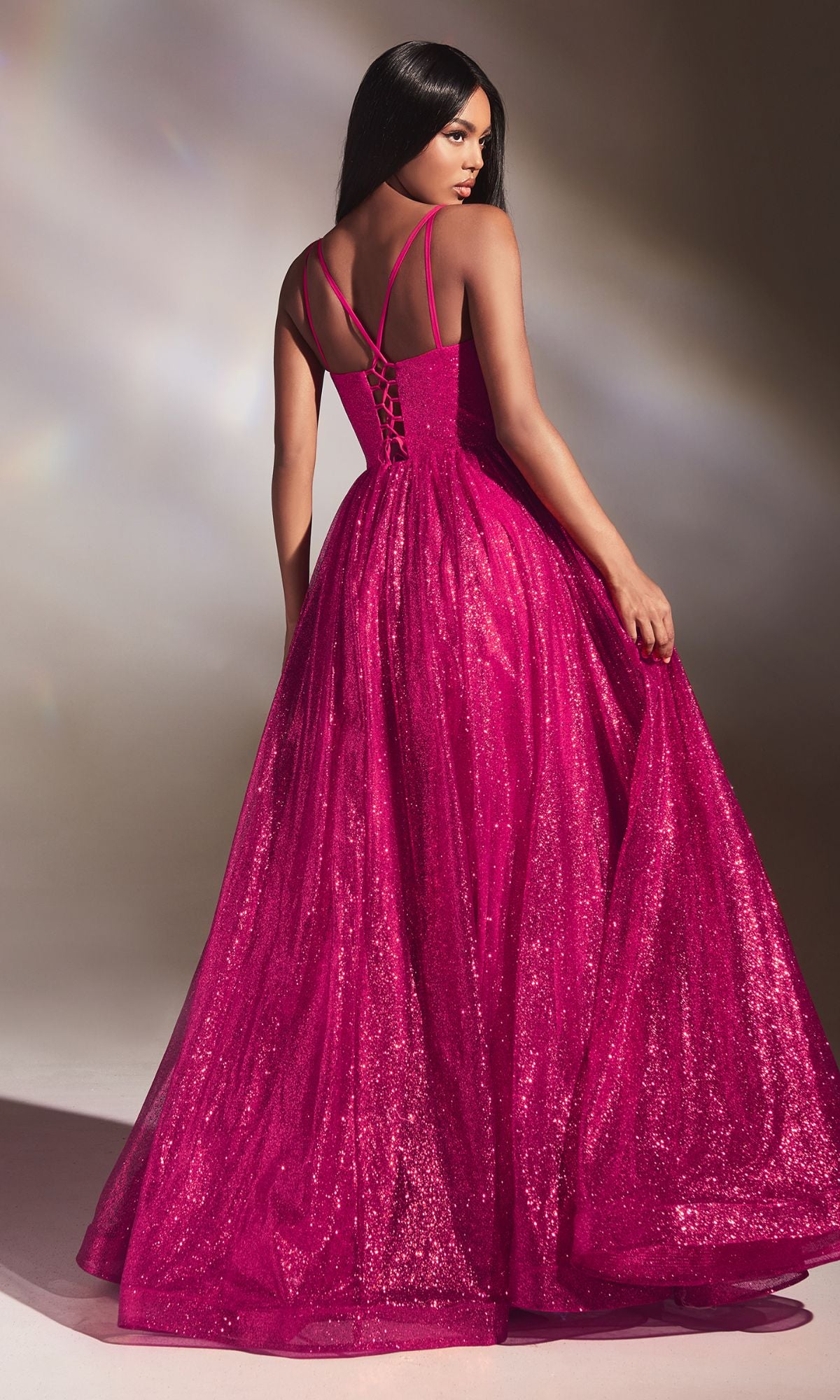 A woman wears the Corset-Back Long Glitter Prom Ball Gown CD996 in bright pink with spaghetti straps and a lace-up back, standing and looking over her shoulder.