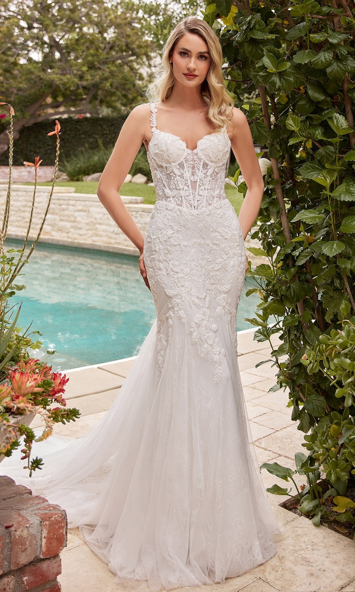 A woman in the Chantilly-Lace Long Off-White Formal Dress CDS432W with a corset waist, sheer bodice, and floral lace details stands by a poolside garden.