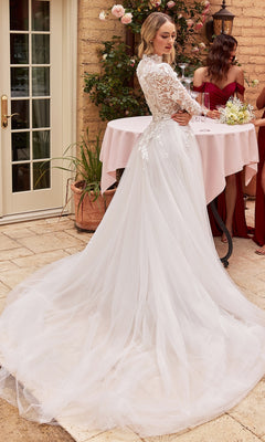 A woman wears the Off-White Long Sleeve Lace Wedding Gown CDS433W outdoors near a round table, her white lace gown glowing, with two women in burgundy dresses in the background.