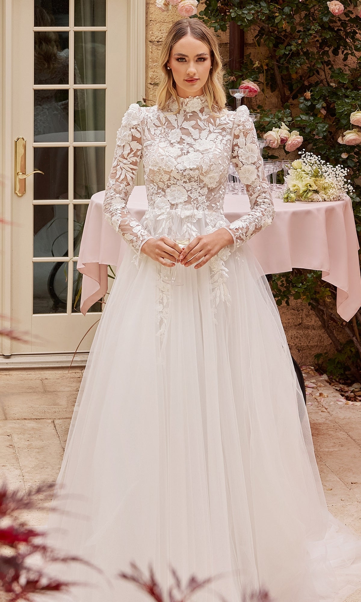 A woman stands outdoors by a table, wearing the Off-White Long Sleeve Lace Wedding Gown CDS433W, featuring floral details and a tulle skirt.