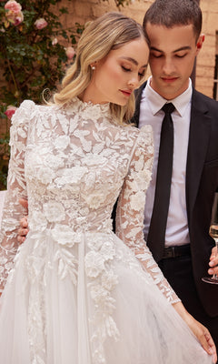 A bride in the Off-White Long Sleeve Lace Wedding Gown CDS433W stands beside a groom in a black suit and tie, holding a glass, with lush greenery in the background.