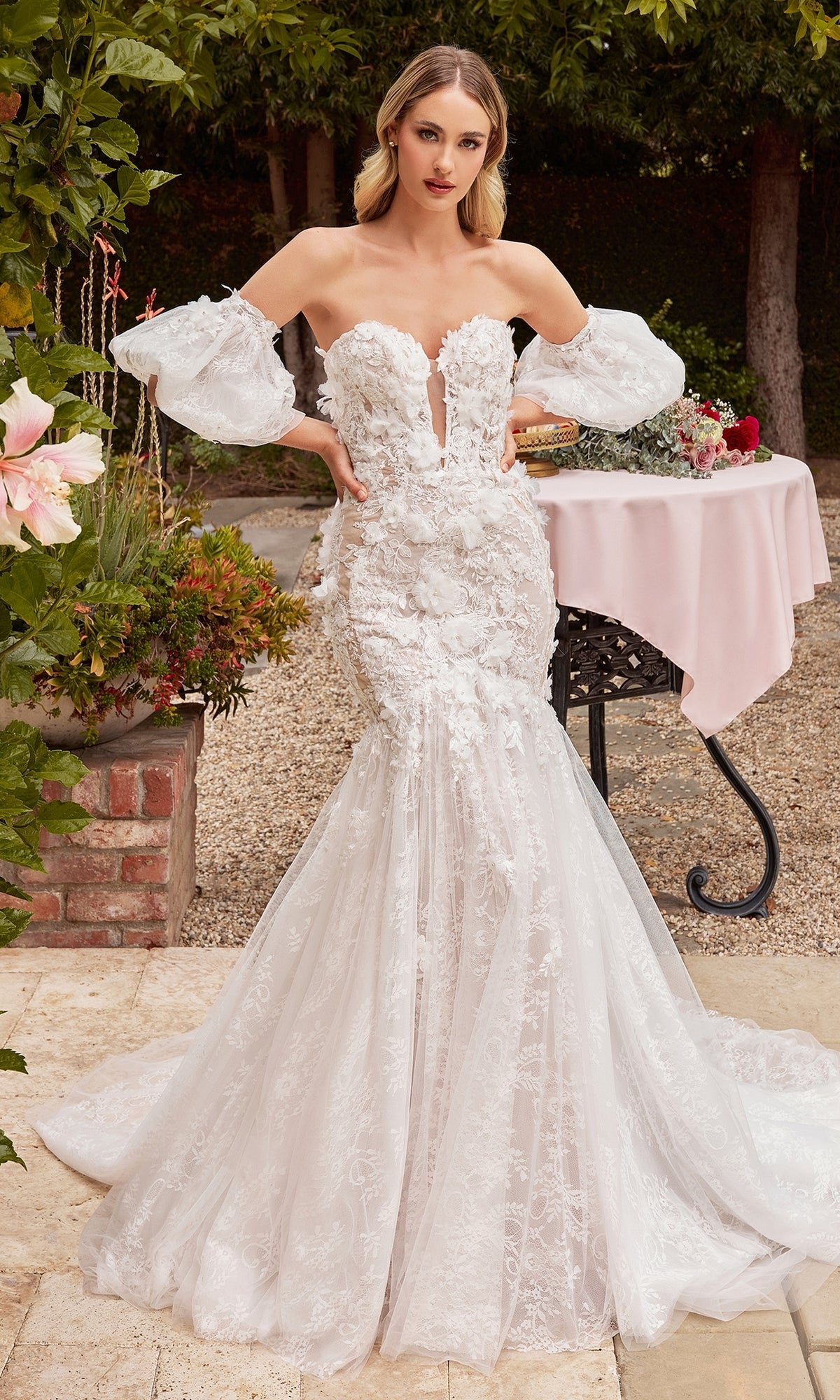 A woman stands outdoors near a table and greenery, wearing the Off-White Long Lace Mermaid Wedding Dress CDS434W with floral appliqué and sheer puff sleeves.