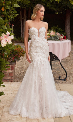 A woman in the Off-White Long Lace Mermaid Wedding Dress CDS434W stands on a stone patio near a flower-adorned table, surrounded by lush greenery.