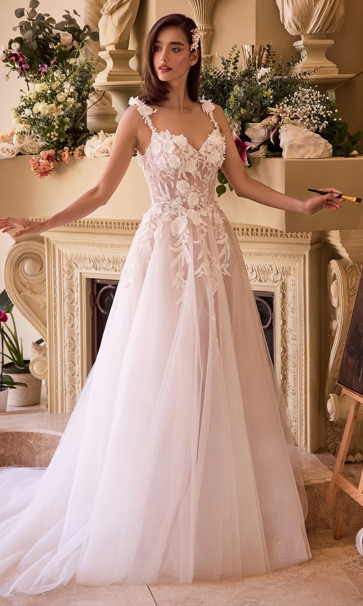 A woman wearing the Long Off-White Bridal Gown: Ladivine CDS498W stands before an ornate, flower-adorned fireplace.