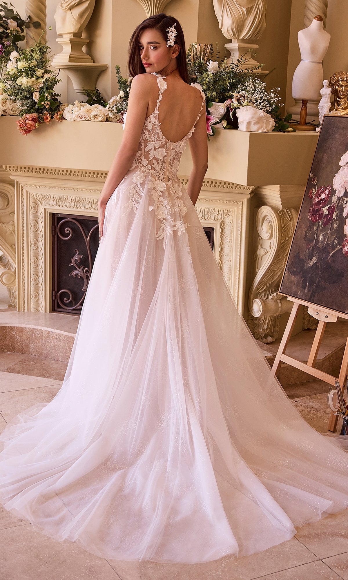 A woman wearing the Ladivine CDS498W long off-white bridal gown of lace and tulle stands indoors near floral arrangements and classical decor, facing away from the camera.