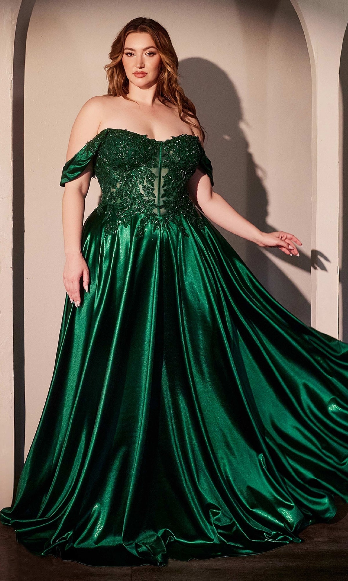 A woman stands gracefully in front of an arched background, wearing the Off-the-Shoulder Prom Ball Gown: Ladivine CDS525—a floor-length emerald green satin dress with a lace bodice.
