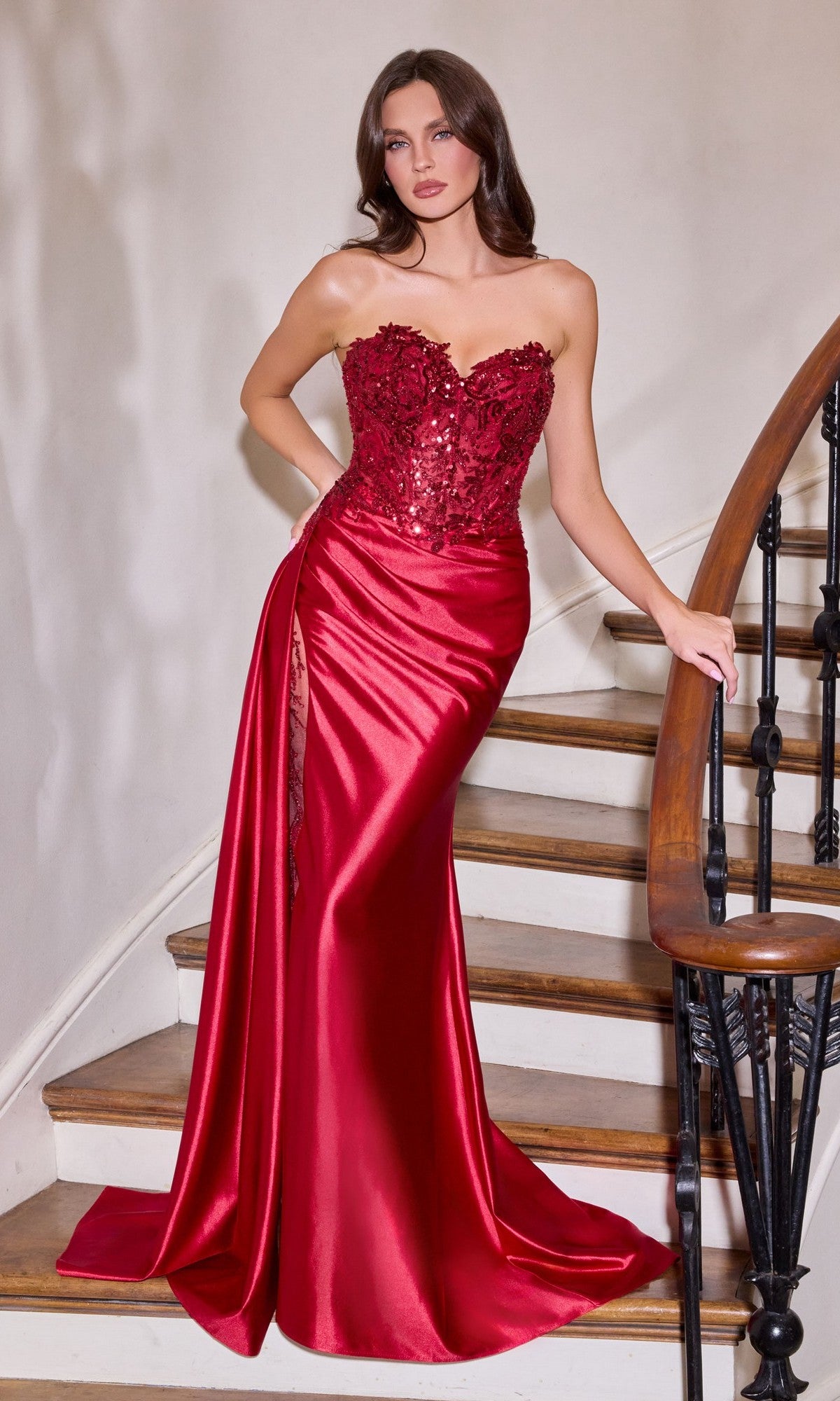 A woman in a burgundy Ladivine CDS565 long prom dress with a sequin corset bodice, strapless neckline, and thigh-high slit stands on a staircase, resting one hand on the railing.