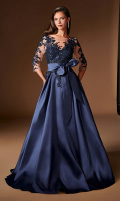 A woman wearing the Ladivine CDS569 Long Formal Dress—a floor-length, dark blue evening gown featuring a lace bodice, sheer sleeves, and a satin bow at the waist—stands against a neutral brown background.
