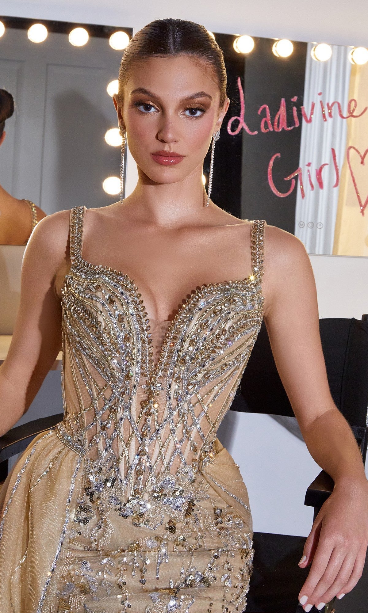 A woman in a gold beaded Ladivine CDS570 long prom dress sits at a lit vanity with 