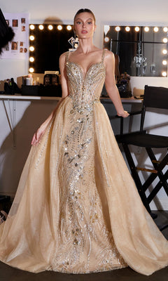 A woman stands in a dressing room wearing the Ladivine CDS570 Long Prom Dress, featuring gold crystal embellishments, a fitted bodice, sheer panels, and flared skirt. Makeup lights and cosmetics in the background complete this formal event look.