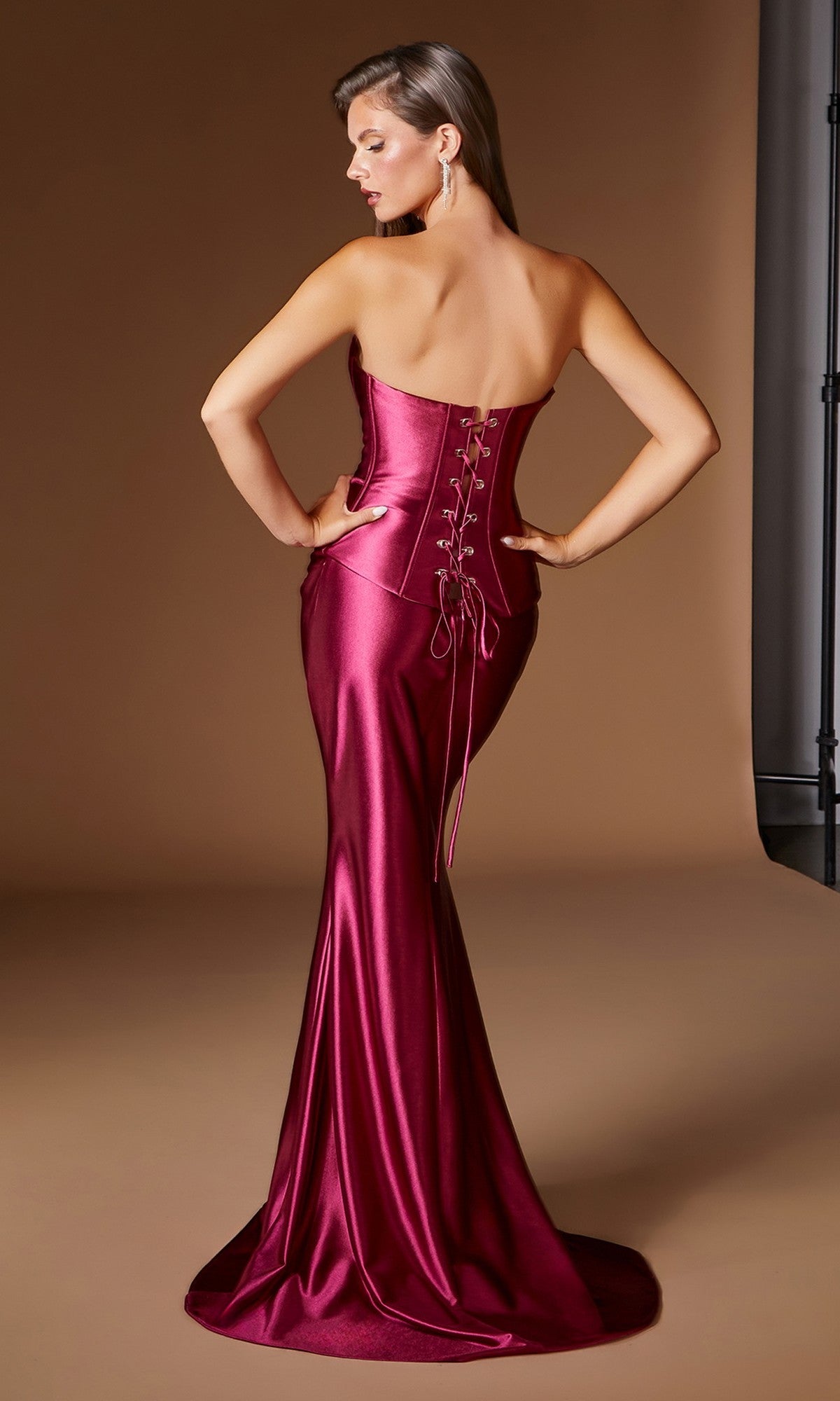 A woman stands facing away against a plain brown background, highlighting the sleek silhouette of the Ladivine CH2602 Long Prom Dress—a shiny, strapless magenta mermaid gown featuring a lace-up corset back.