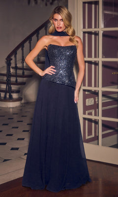 A woman stands indoors in front of glass doors and a staircase, wearing the Ladivine CH2603 long prom dress in navy, which features a strapless embellished corset bodice and a floor-length sequin skirt.