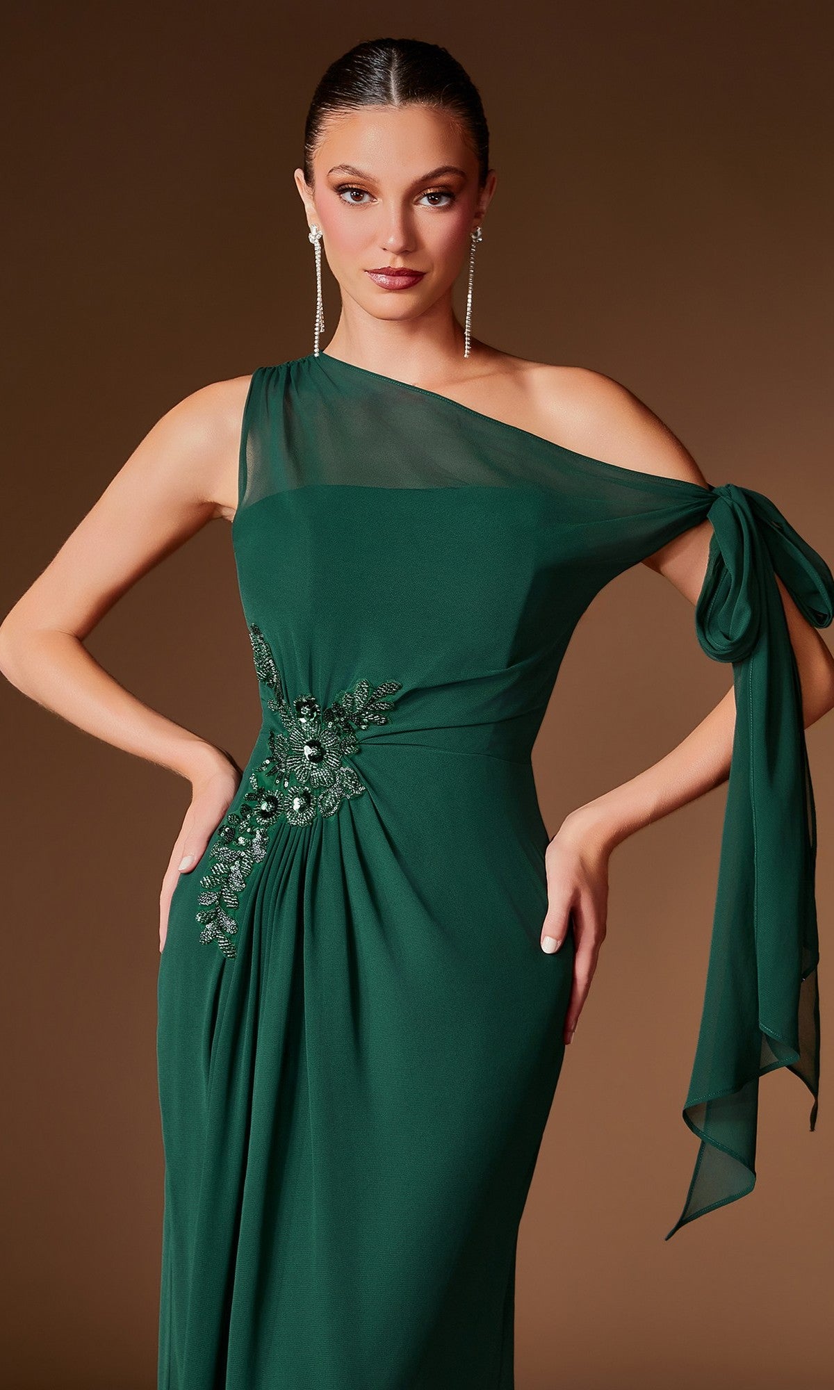 A woman models the Ladivine CH2612 long formal dress—an elegant, one-shoulder emerald green gown featuring beaded floral embellishments and a bow detail—against a plain brown background.