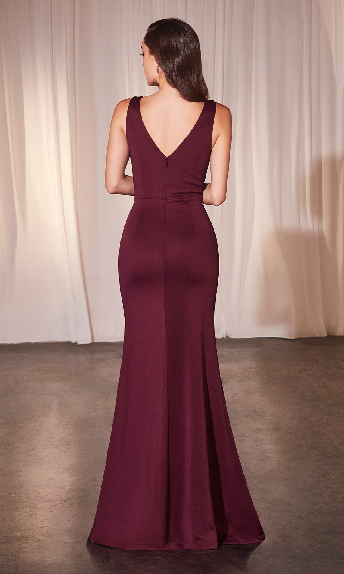 A woman faces away from the camera in a sleeveless, floor-length Ladivine CH377 Drape-Front Long Prom Dress in plus size burgundy with a deep V-back. Cream-colored curtains hang in the background.