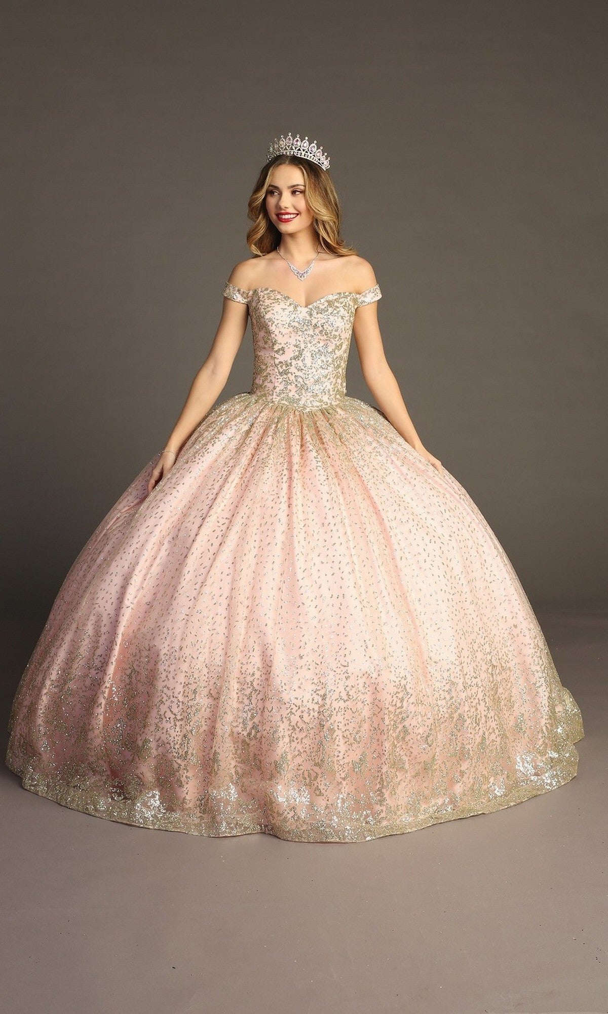 A woman in the Quinceanera Dress CHF3001 By Chicas, featuring a sparkling blush hue with metallic embroidery and a tiara, stands against a plain gray background, smiling with her arms slightly out to the side.