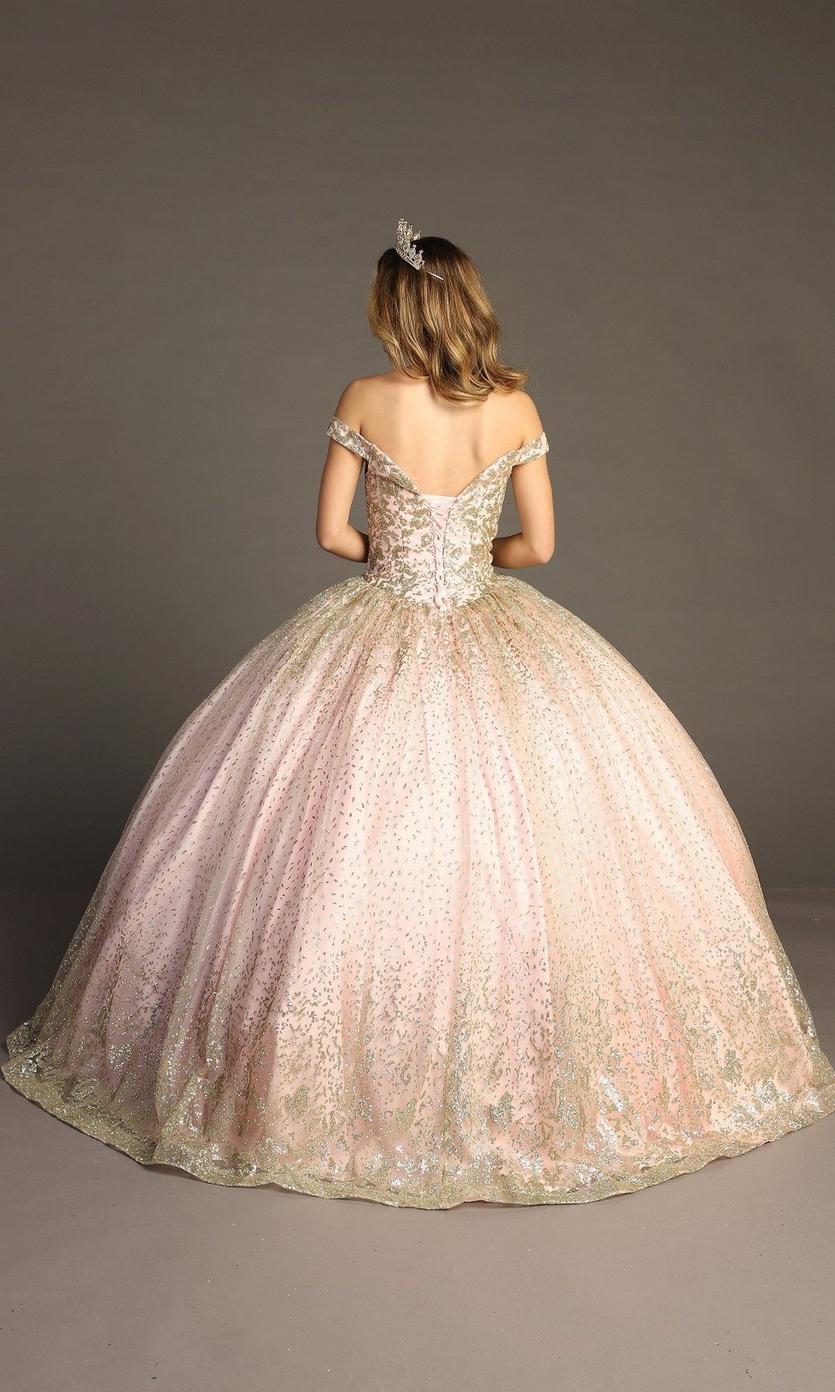 A person wearing the Quinceanera Dress CHF3001 By Chicas—an off-the-shoulder pink gown with a voluminous ballgown skirt and delicate metallic embroidery—stands facing away against a plain background, crowned with a sparkling tiara.