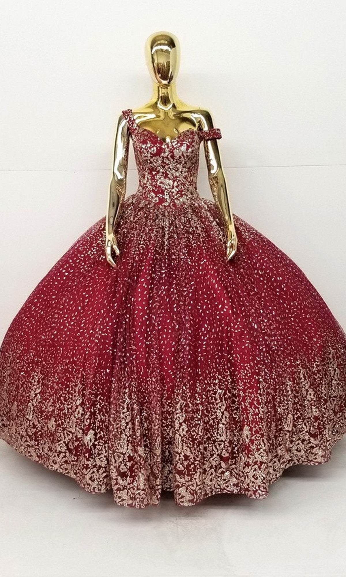 A gold mannequin displays the Quinceanera Dress CHF3001 by Chicas, featuring an off-shoulder, sparkling burgundy ball gown with a voluminous skirt and intricate metallic embroidery, set against a plain white background.