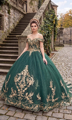 A woman in the Quinceanera Dress CHF3006 By Chicas, featuring green fabric with gold embroidery and an ornate off-the-shoulder design, stands on a cobblestone path near stone stairs and ivy-covered walls.