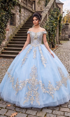 A woman wearing the Quinceanera Dress CHF3006 By Chicas in baby blue and gold stands on a cobblestone path beside stone steps and ivy-covered walls.