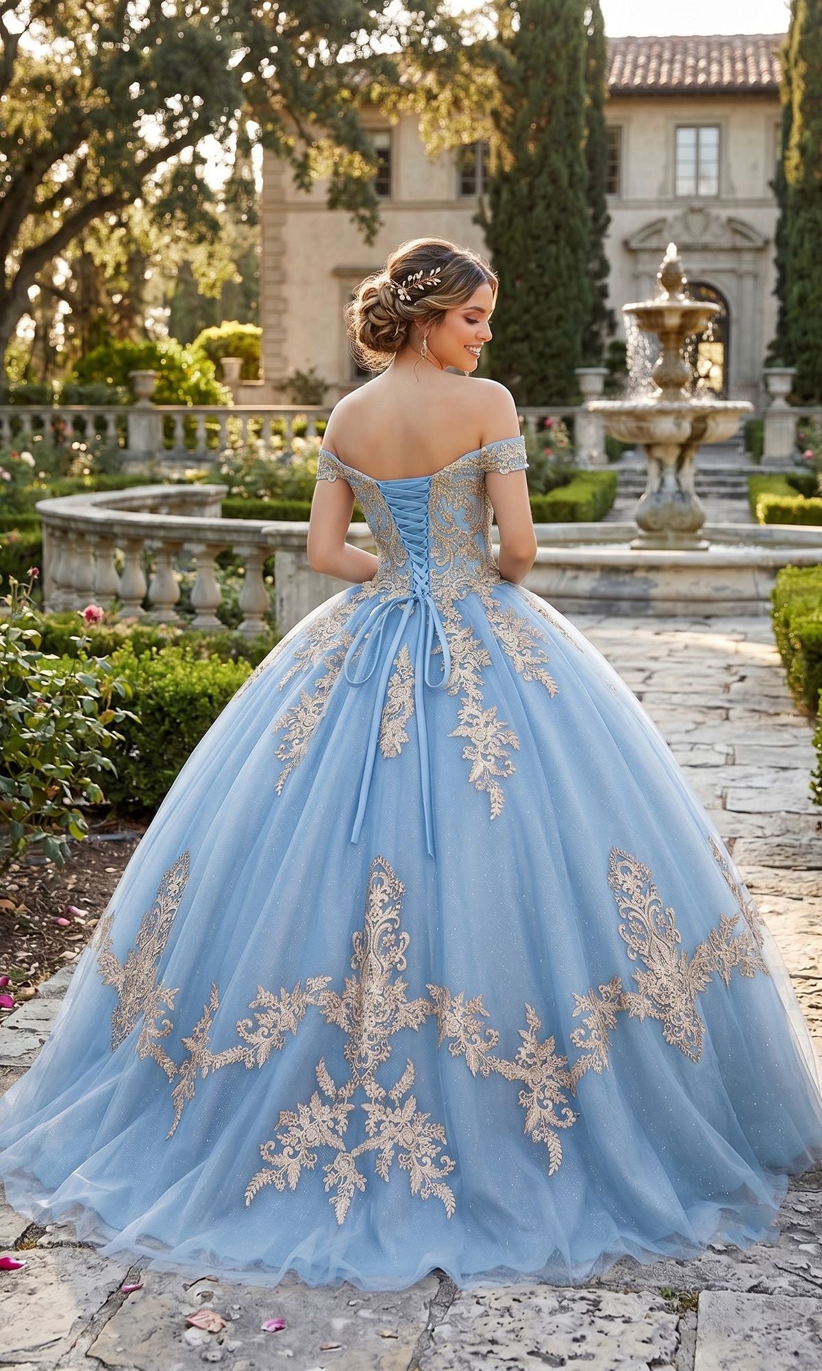 Wearing the Quinceanera Dress CHF3008 By Chicas—a stunning blue gown adorned with gold embroidery—a woman stands on a stone path in a formal garden, her back turned to reveal a fountain and grand house in the background.