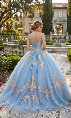 Wearing the Quinceanera Dress CHF3008 By Chicas—a stunning blue gown adorned with gold embroidery—a woman stands on a stone path in a formal garden, her back turned to reveal a fountain and grand house in the background.