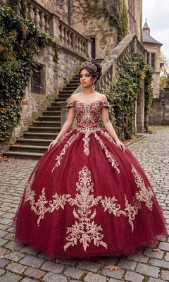 Wearing the Quinceanera Dress CHF3008 By Chicas in burgundy and gold, a woman with a crown stands on a cobblestone path before ivy-covered stone stairs and an old building.