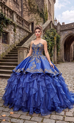 A woman in the Quinceanera Dress CHF3009 By Chicas, featuring royal blue and gold embroidery with a voluminous ruffled organza skirt, stands on a cobblestone path near a historic stone building and steps.