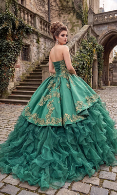 A woman wearing the Quinceanera Dress CHF3009 By Chicas, a green strapless gown with gold embroidery, stands on a cobblestone path near stone stairs and ivy-covered walls, glancing over her shoulder. The satin bodice and ruffled organza skirt of her dress catch the soft light, creating an elegant and enchanting silhouette.