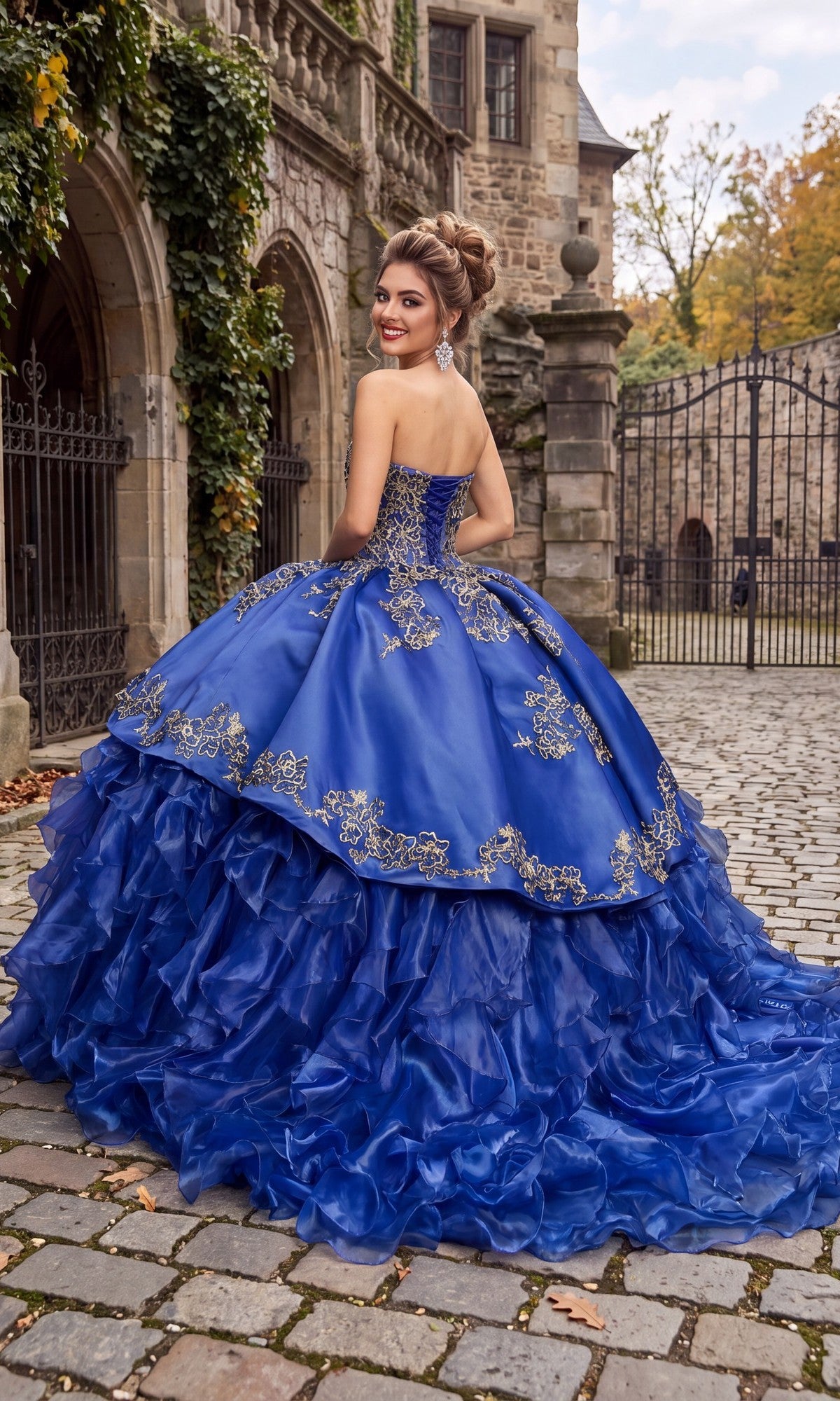 A woman wearing the Quinceanera Dress CHF3009 by Chicas—with its strapless satin bodice adorned in gold embroidery—stands on a cobblestone path outside a stone building, facing away and smiling over her shoulder as the gown’s ruffled organza skirt flows elegantly behind her.